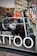 A motorcycle is displayed inside a shop with a large window. The bike features a bold orange fuel tank and is equipped with knobby tires, indicating off-road capabilities. Reflections on the glass suggest a street setting, and visible signage on the window includes the word 'TATTOO' in large white letters.