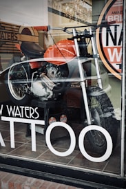 A motorcycle is displayed inside a shop with a large window. The bike features a bold orange fuel tank and is equipped with knobby tires, indicating off-road capabilities. Reflections on the glass suggest a street setting, and visible signage on the window includes the word 'TATTOO' in large white letters.