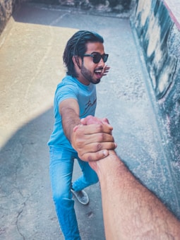 A man wearing sunglasses and a light blue t-shirt is reaching out and holding another person's hand in a firm grip. He is standing outdoors, with a wall visible in the background. The perspective suggests that the photo is taken from a high angle, focusing on the handshake.