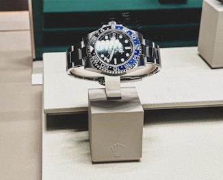 A sleek, luxurious wristwatch with a rotating bezel and a prominent metallic bracelet is displayed on a cushioned stand. The watch face has multiple time indicators, luminescent hands, and a deep blue and black color scheme. The setting appears to be a high-end retail store with elegant packaging and branding.