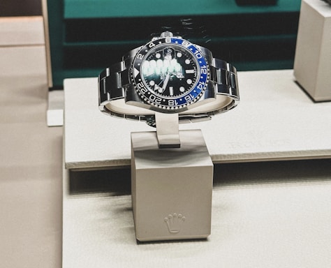 A sleek, luxurious wristwatch with a rotating bezel and a prominent metallic bracelet is displayed on a cushioned stand. The watch face has multiple time indicators, luminescent hands, and a deep blue and black color scheme. The setting appears to be a high-end retail store with elegant packaging and branding.