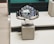 A sleek, luxurious wristwatch with a rotating bezel and a prominent metallic bracelet is displayed on a cushioned stand. The watch face has multiple time indicators, luminescent hands, and a deep blue and black color scheme. The setting appears to be a high-end retail store with elegant packaging and branding.