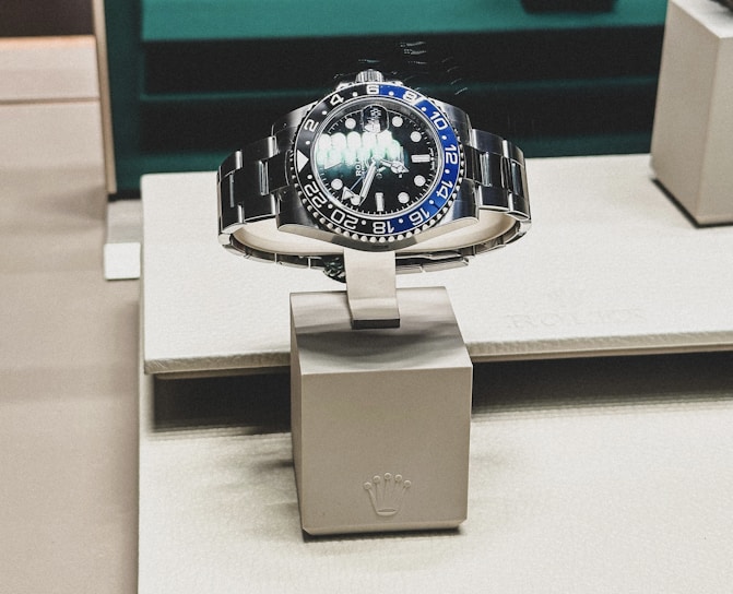 A sleek, luxurious wristwatch with a rotating bezel and a prominent metallic bracelet is displayed on a cushioned stand. The watch face has multiple time indicators, luminescent hands, and a deep blue and black color scheme. The setting appears to be a high-end retail store with elegant packaging and branding.