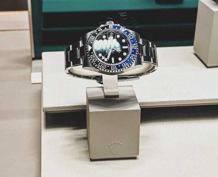 A sleek, luxurious wristwatch with a rotating bezel and a prominent metallic bracelet is displayed on a cushioned stand. The watch face has multiple time indicators, luminescent hands, and a deep blue and black color scheme. The setting appears to be a high-end retail store with elegant packaging and branding.