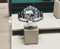 A sleek, luxurious wristwatch with a rotating bezel and a prominent metallic bracelet is displayed on a cushioned stand. The watch face has multiple time indicators, luminescent hands, and a deep blue and black color scheme. The setting appears to be a high-end retail store with elegant packaging and branding.