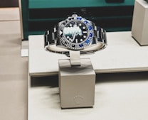 A sleek, luxurious wristwatch with a rotating bezel and a prominent metallic bracelet is displayed on a cushioned stand. The watch face has multiple time indicators, luminescent hands, and a deep blue and black color scheme. The setting appears to be a high-end retail store with elegant packaging and branding.