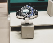 A sleek, luxurious wristwatch with a rotating bezel and a prominent metallic bracelet is displayed on a cushioned stand. The watch face has multiple time indicators, luminescent hands, and a deep blue and black color scheme. The setting appears to be a high-end retail store with elegant packaging and branding.