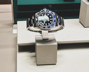 A sleek, luxurious wristwatch with a rotating bezel and a prominent metallic bracelet is displayed on a cushioned stand. The watch face has multiple time indicators, luminescent hands, and a deep blue and black color scheme. The setting appears to be a high-end retail store with elegant packaging and branding.