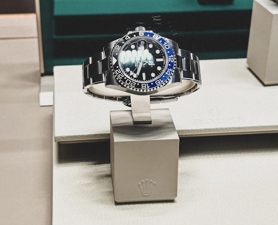 A sleek, luxurious wristwatch with a rotating bezel and a prominent metallic bracelet is displayed on a cushioned stand. The watch face has multiple time indicators, luminescent hands, and a deep blue and black color scheme. The setting appears to be a high-end retail store with elegant packaging and branding.