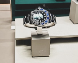 A sleek, luxurious wristwatch with a rotating bezel and a prominent metallic bracelet is displayed on a cushioned stand. The watch face has multiple time indicators, luminescent hands, and a deep blue and black color scheme. The setting appears to be a high-end retail store with elegant packaging and branding.