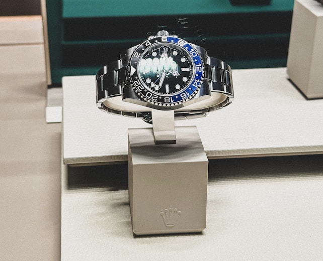 A sleek, luxurious wristwatch with a rotating bezel and a prominent metallic bracelet is displayed on a cushioned stand. The watch face has multiple time indicators, luminescent hands, and a deep blue and black color scheme. The setting appears to be a high-end retail store with elegant packaging and branding.