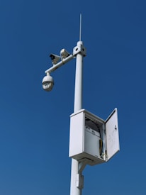 A tall pole equipped with a surveillance camera and an open electrical box against a clear blue sky. The camera housing appears spherical, and the electrical box reveals wiring and components inside.