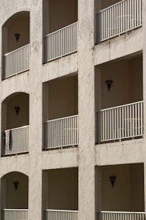 A building facade featuring multiple balconies with metal railings and recessed walls in a neutral tone. Each balcony has a triangular wall sconce, and one has a hanging towel. The walls show signs of weathering.