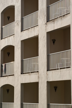 A building facade featuring multiple balconies with metal railings and recessed walls in a neutral tone. Each balcony has a triangular wall sconce, and one has a hanging towel. The walls show signs of weathering.