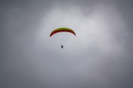 A paraglider is soaring through a cloudy sky, featuring a bright green and red wing. The silhouette of the pilot is visible beneath the canopy, suspended and navigating the air currents.