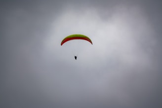 A paraglider is soaring through a cloudy sky, featuring a bright green and red wing. The silhouette of the pilot is visible beneath the canopy, suspended and navigating the air currents.