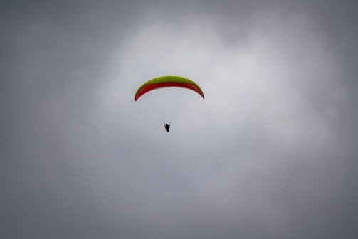A paraglider is soaring through a cloudy sky, featuring a bright green and red wing. The silhouette of the pilot is visible beneath the canopy, suspended and navigating the air currents.