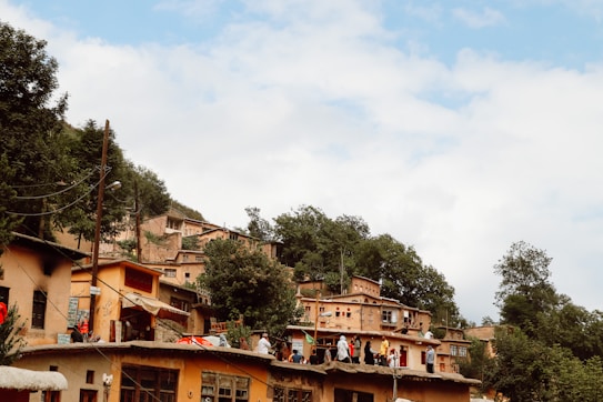 A hillside village with houses made from earthy materials and interconnected roofs. Trees and greenery surround the area, enhancing the rustic and traditional charm. Several people are gathered on one of the flat rooftops, engaging in activity or conversation. The sky is partially cloudy, contributing to a serene atmosphere.