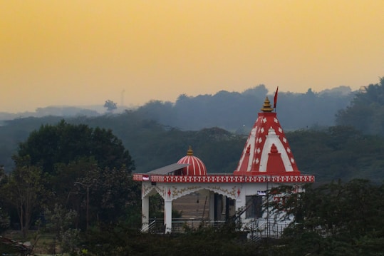A temple with a red and white color scheme is situated amidst lush greenery. The temple features intricate designs and a conical dome with a golden pinnacle. The background shows a hazy sky with fading daylight, casting a serene and peaceful ambiance.