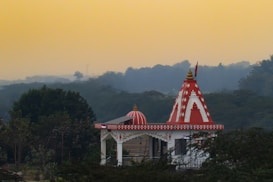 A temple with a red and white color scheme is situated amidst lush greenery. The temple features intricate designs and a conical dome with a golden pinnacle. The background shows a hazy sky with fading daylight, casting a serene and peaceful ambiance.