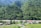 A lush green mountainous region with dense foliage covering the slopes. A river winds its way through the valley, with small buildings and houses scattered near its banks. The scene is rural and tranquil, with clear skies and abundant greenery.
