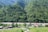 A lush green mountainous region with dense foliage covering the slopes. A river winds its way through the valley, with small buildings and houses scattered near its banks. The scene is rural and tranquil, with clear skies and abundant greenery.