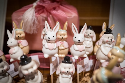 A collection of ceramic bunny figurines, with some wearing pink bows or black hats, arranged on sticks. They are surrounded by a soft pink packaging, creating a festive and whimsical display.