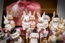 A collection of ceramic bunny figurines, with some wearing pink bows or black hats, arranged on sticks. They are surrounded by a soft pink packaging, creating a festive and whimsical display.