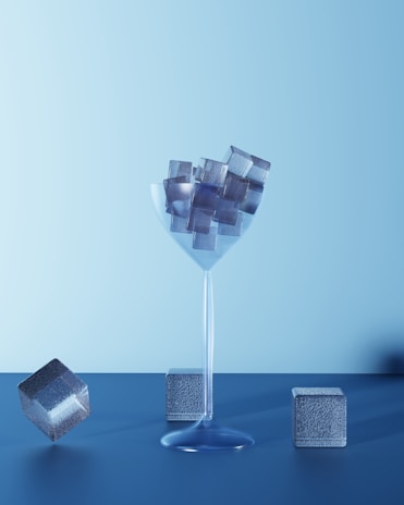 A transparent glass filled with metallic cubes, placed on a blue surface with a soft gradient blue background. Several cubes are positioned around the glass, some lying flat and one slightly elevated.