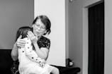 An elderly woman with glasses embraces a young girl wearing a polka-dotted dress. They are seated on a dark-colored sofa in a room with minimal decorations. The woman appears to be comforting the child, gently holding her face.
