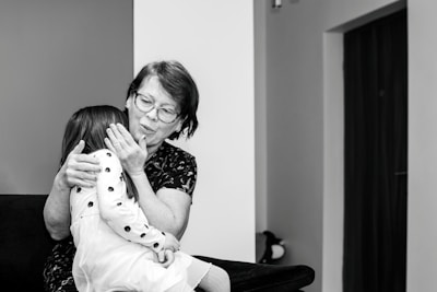 An elderly woman with glasses embraces a young girl wearing a polka-dotted dress. They are seated on a dark-colored sofa in a room with minimal decorations. The woman appears to be comforting the child, gently holding her face.
