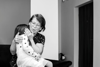 An elderly woman with glasses embraces a young girl wearing a polka-dotted dress. They are seated on a dark-colored sofa in a room with minimal decorations. The woman appears to be comforting the child, gently holding her face.