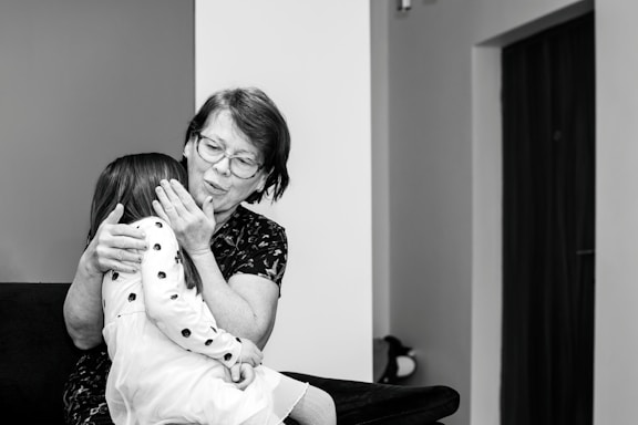 An elderly woman with glasses embraces a young girl wearing a polka-dotted dress. They are seated on a dark-colored sofa in a room with minimal decorations. The woman appears to be comforting the child, gently holding her face.