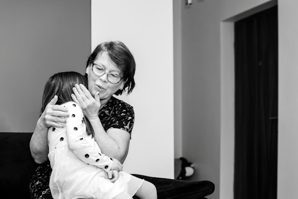 An elderly woman with glasses embraces a young girl wearing a polka-dotted dress. They are seated on a dark-colored sofa in a room with minimal decorations. The woman appears to be comforting the child, gently holding her face.