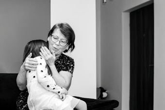An elderly woman with glasses embraces a young girl wearing a polka-dotted dress. They are seated on a dark-colored sofa in a room with minimal decorations. The woman appears to be comforting the child, gently holding her face.