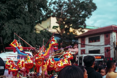 Colorful dragon puppets with intricate designs and vibrant colors are displayed prominently. A group of people is gathered, likely at a festive outdoor event with a building and large trees in the background.