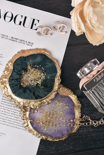A stylish composition featuring a Vogue magazine page, elegant earrings with a recognizable logo, two decorative coasters with gold edging and gold flakes, a floral arrangement, and a perfume bottle. The scene is set on a dark wooden surface, creating a chic and luxurious aesthetic.