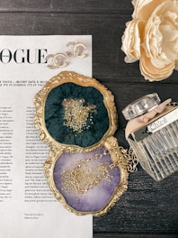 A flat lay image featuring a stylish arrangement on a dark wooden surface. There is a decorative magazine page from Vogue, elegant Chanel earrings with the brand's iconic logo, and a Chloe perfume bottle with a ribbon. Two agate slices with gold-colored edges are used as coasters; one is a deep green with gold flecks and the other is a light purple with a gold chain placed on it. A cream-colored peony flower in full bloom also adds a sophisticated touch.