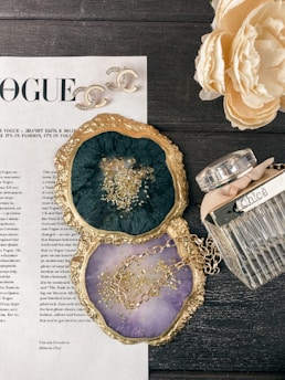 A flat lay image featuring a stylish arrangement on a dark wooden surface. There is a decorative magazine page from Vogue, elegant Chanel earrings with the brand's iconic logo, and a Chloe perfume bottle with a ribbon. Two agate slices with gold-colored edges are used as coasters; one is a deep green with gold flecks and the other is a light purple with a gold chain placed on it. A cream-colored peony flower in full bloom also adds a sophisticated touch.