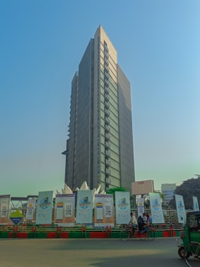 A tall, modern skyscraper with a rectangular design stands prominently against a clear blue sky. Below the building, there are a series of large advertisement boards along a street. Several people are visible, including some on bicycles, and a green auto-rickshaw is also present.