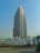 A tall, modern skyscraper with a rectangular design stands prominently against a clear blue sky. Below the building, there are a series of large advertisement boards along a street. Several people are visible, including some on bicycles, and a green auto-rickshaw is also present.
