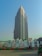A tall, modern skyscraper with a rectangular design stands prominently against a clear blue sky. Below the building, there are a series of large advertisement boards along a street. Several people are visible, including some on bicycles, and a green auto-rickshaw is also present.