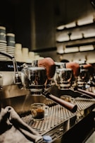 A close-up of a row of espresso machines with portafilters at a café, with freshly brewed espresso in transparent cups underneath. The machines have a polished metal finish and wooden handles, reflecting soft ambient lighting.