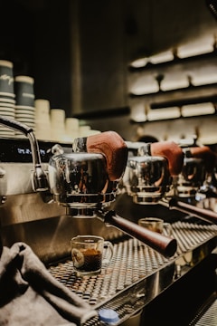 A close-up of a row of espresso machines with portafilters at a café, with freshly brewed espresso in transparent cups underneath. The machines have a polished metal finish and wooden handles, reflecting soft ambient lighting.