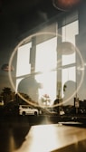 A reflective scene combining indoor lighting fixtures and an outdoor urban landscape. Large windows allow sunlight to enter, casting shadows and reflections on the glass. A circular light effect frames the scene, possibly from the lens or lighting. Vehicles are visible outside along with palm trees, indicating a city or coastal environment.