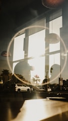 A reflective scene combining indoor lighting fixtures and an outdoor urban landscape. Large windows allow sunlight to enter, casting shadows and reflections on the glass. A circular light effect frames the scene, possibly from the lens or lighting. Vehicles are visible outside along with palm trees, indicating a city or coastal environment.