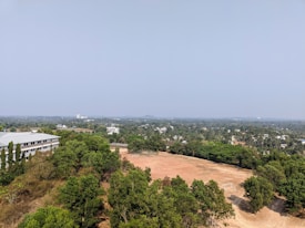 Aerial view of a landscape featuring a sports field surrounded by dense green vegetation. A building with a white rooftop is visible to the left, while a distant view of a cityscape extends towards the horizon under a clear blue sky.