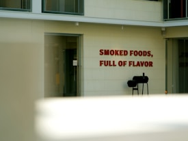 A building exterior displays large, bold red text with the phrase 'Smoked Foods, Full of Flavor'. Adjacent to the wall, a black barbecue smoker stands on the concrete ground. The setting appears modern, with glass doors and windows integrated into the facade.