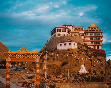 A large, multi-story Buddhist monastery with intricate architectural details sits atop a hill. Below, there are colorful prayer flags strung between poles. An ornate traditional gate stands in the foreground, and piles of logs and rocky terrain surround the area. The sky is partly cloudy, with a mix of blue and white colors.