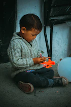 A young child sits cross-legged on the ground, engrossed in playing with a small, colorful toy. The child wears a gray knitted sweater and dark pants, with slightly worn shoes. A bright orange toy lies nearby, and a white balloon rests on the ground next to the child. The background shows a textured wall and a folded chair.